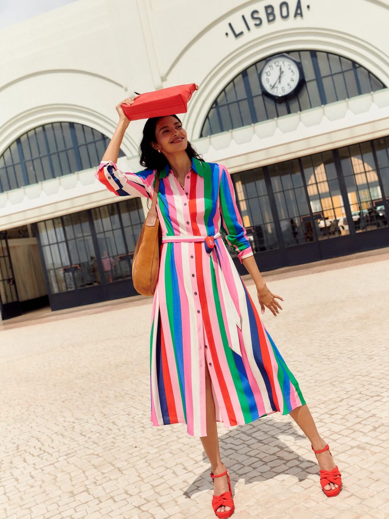 Woman in a colorful striped dress and red shoes holds a red hat above her head, standing outside a building marked "Lisboa.