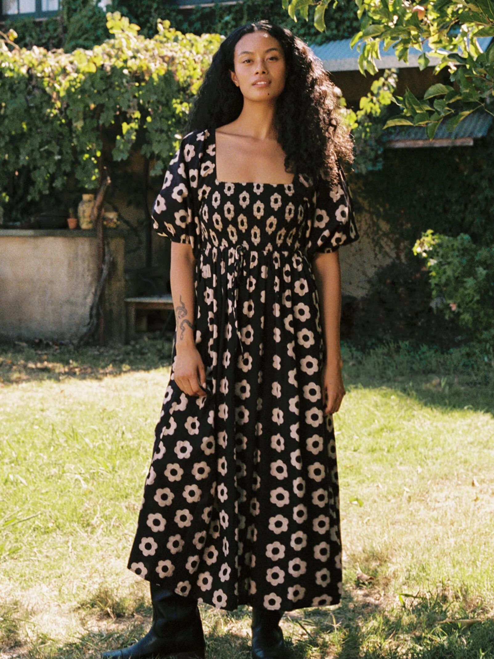 A person in a floral dress stands on grass in a sunlit garden, with trees and a building in the background.