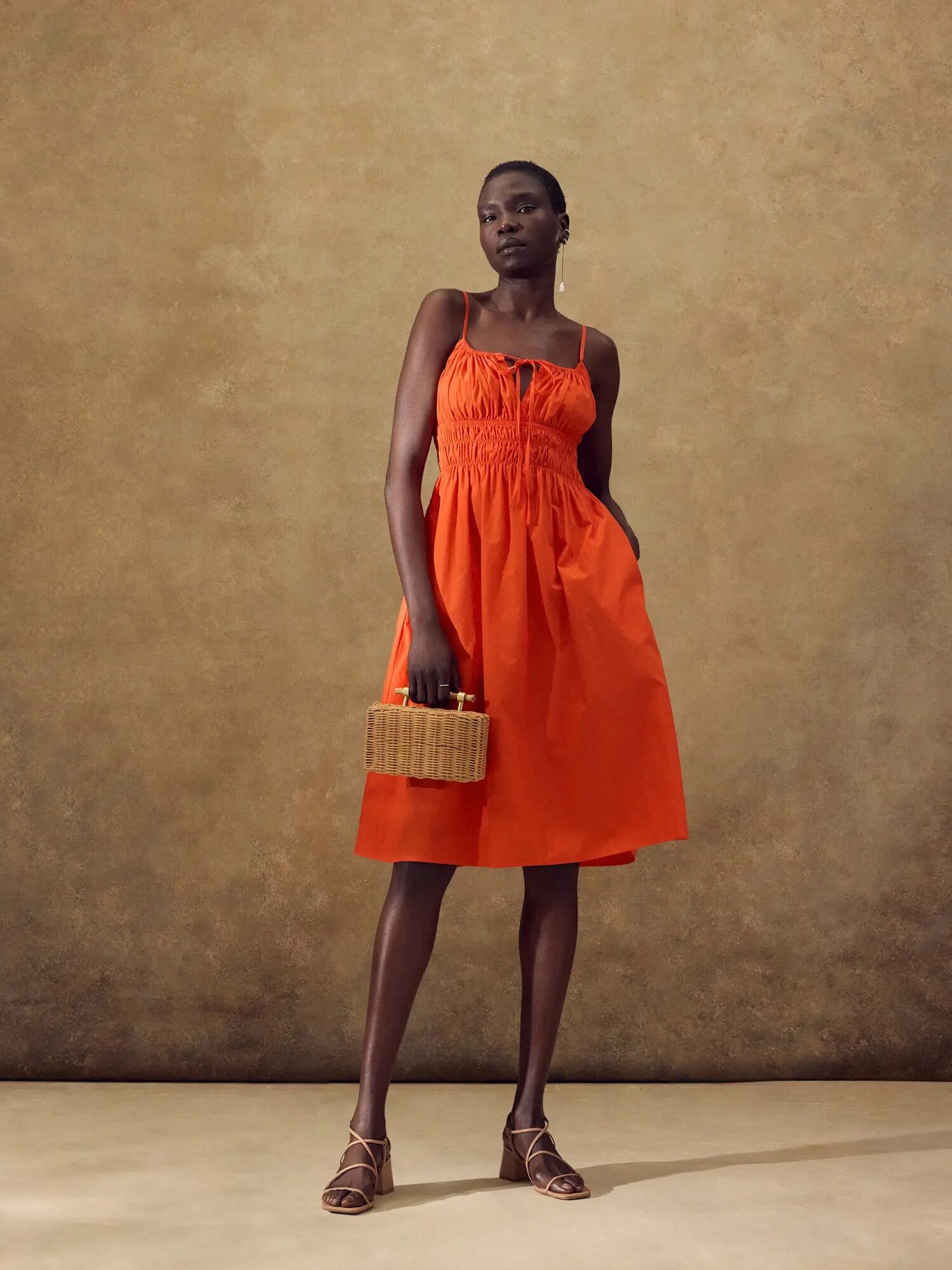 A person stands against a beige backdrop, wearing an orange dress, holding a wicker handbag, and dressed in strappy sandals.