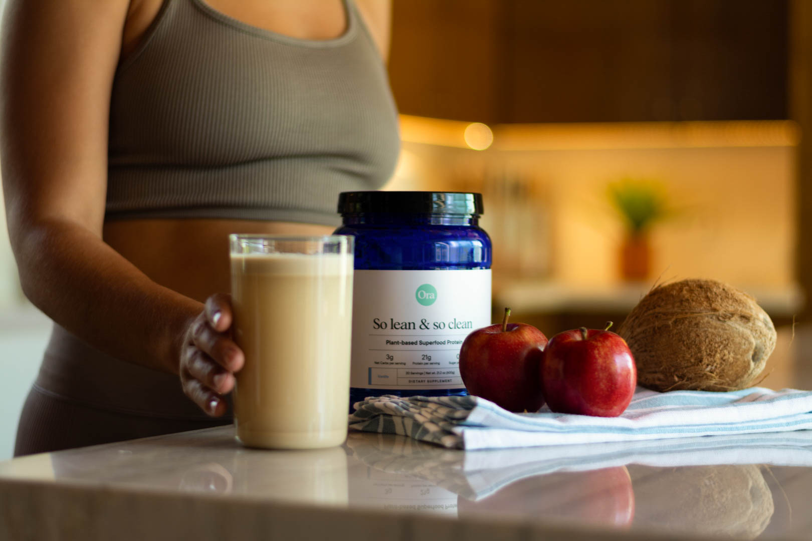 A person holding a glass of a beige drink next to a blue container labeled "So lean & so clean," two apples, and a coconut on a kitchen counter.