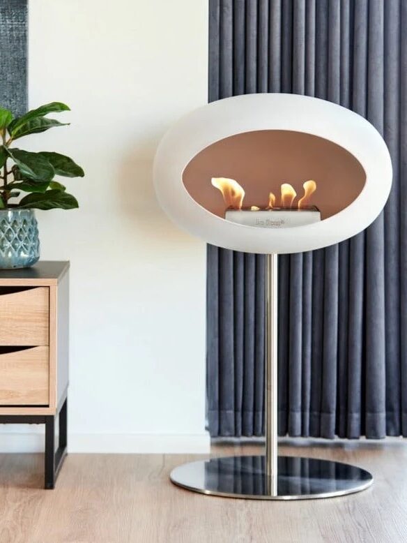 Modern indoor fireplace with a white oval design on a metal stand, near a wooden cabinet with a potted plant, in a room with a window and dark curtains.