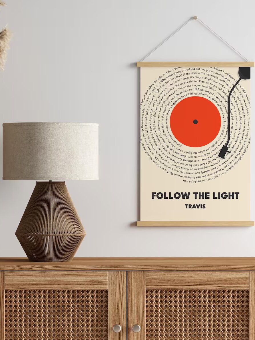 A wooden sideboard with a textured lamp and pampas grass is against a wall. A poster with a record design above text reading "Follow the Light" is hung on the wall.