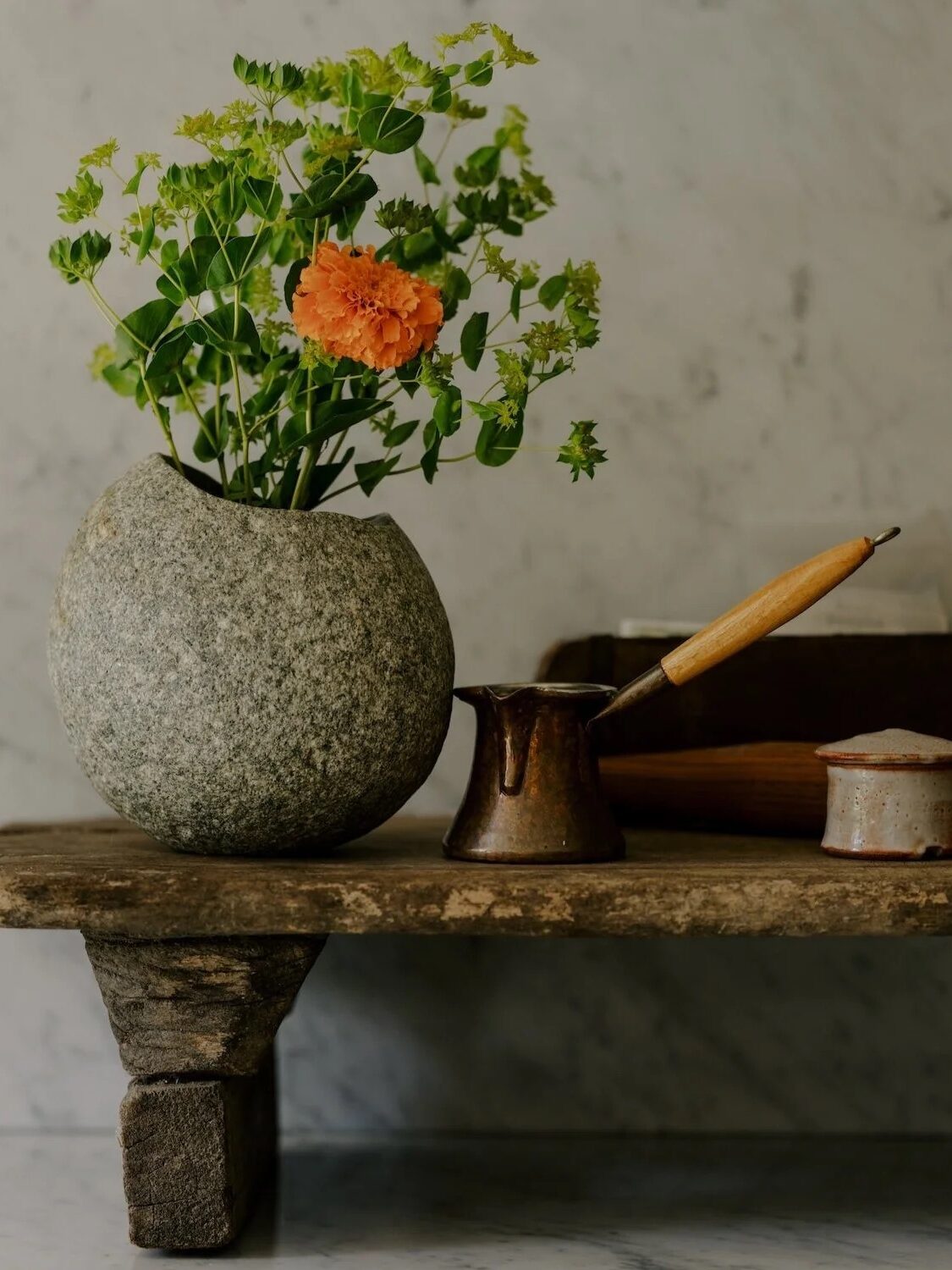 A round stone vase holds greenery and an orange flower on a wooden shelf. Nearby are a small copper pot and a lidded ceramic container.