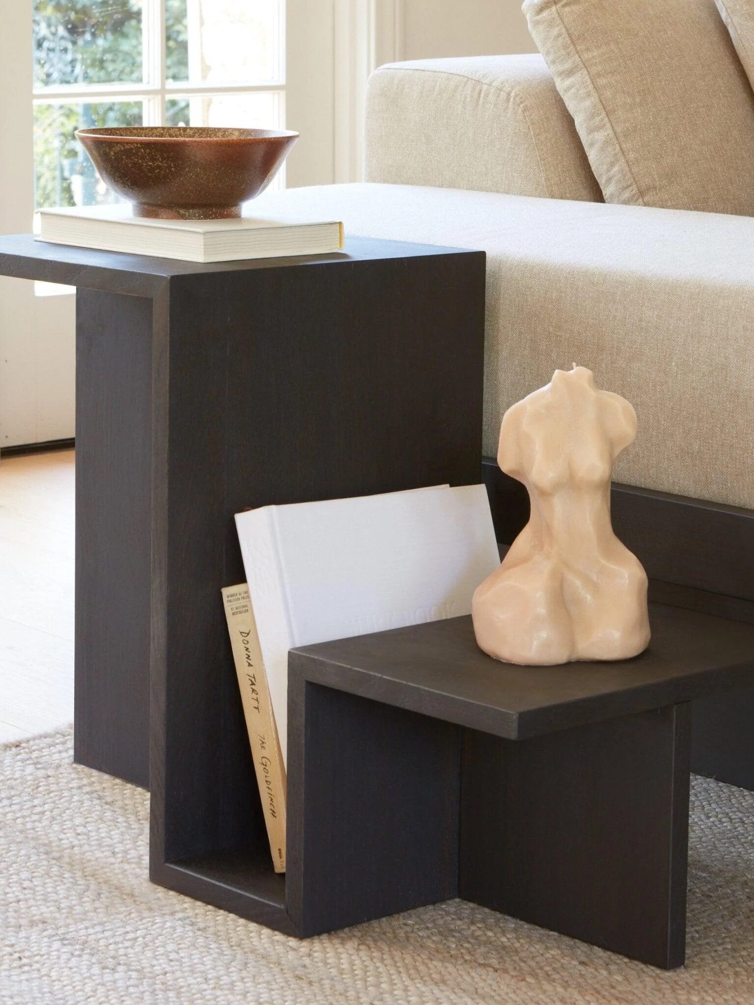 Wooden side table with a geometric design, featuring a ceramic torso sculpture, a bowl, and a book. It is positioned next to a beige sofa on a woven rug.