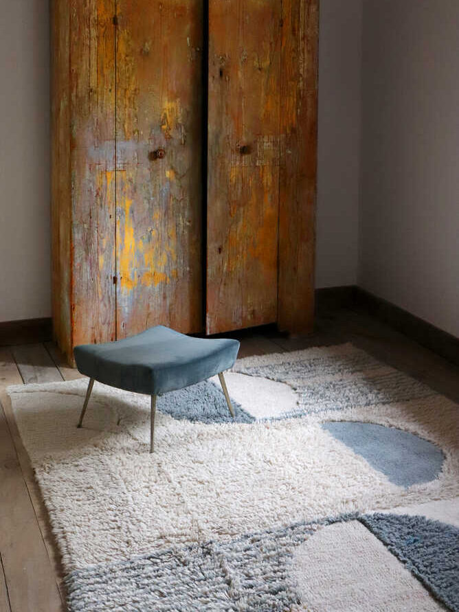 A rustic wooden wardrobe stands against a wall in a room with a textured rug and a small, modern stool.