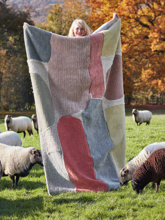 A person holds up a large, colorful patchwork blanket in a grassy field surrounded by sheep and autumn trees.