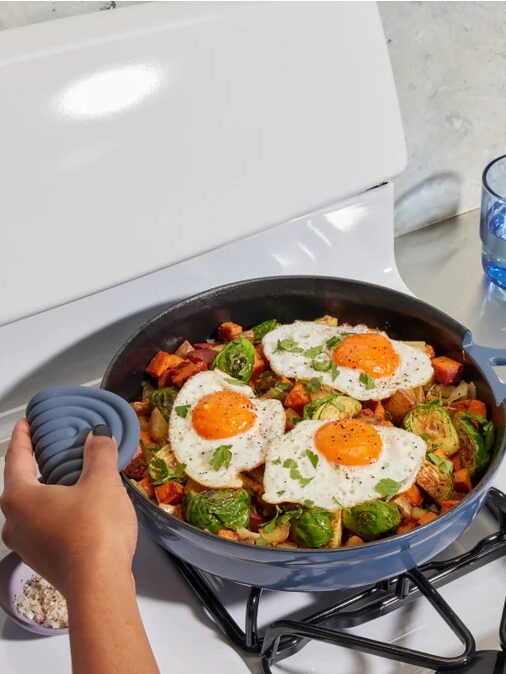 A person holds a skillet on a stove containing fried eggs, Brussels sprouts, and diced potatoes. A glass of water is in the background.