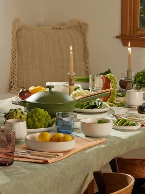 A dining table is set with dishes, candles, vegetables, lemons, and a green pot. The table is surrounded by wooden chairs, and a window lets in natural light.