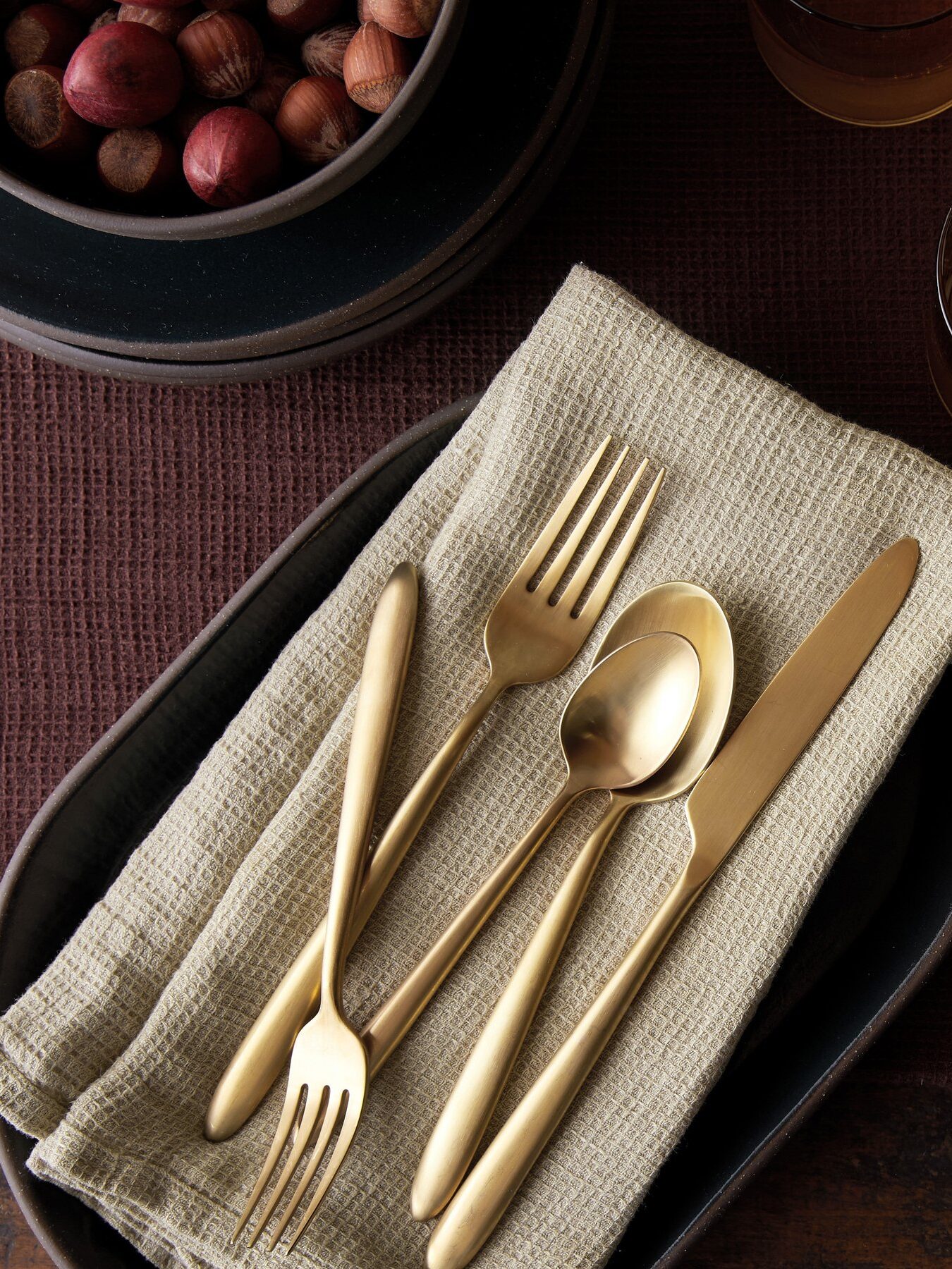 Table setting with gold cutlery on a beige napkin, brown plates, a bowl of nuts, glasses with amber liquid, and a pink granite candle holder on a brown tablecloth.