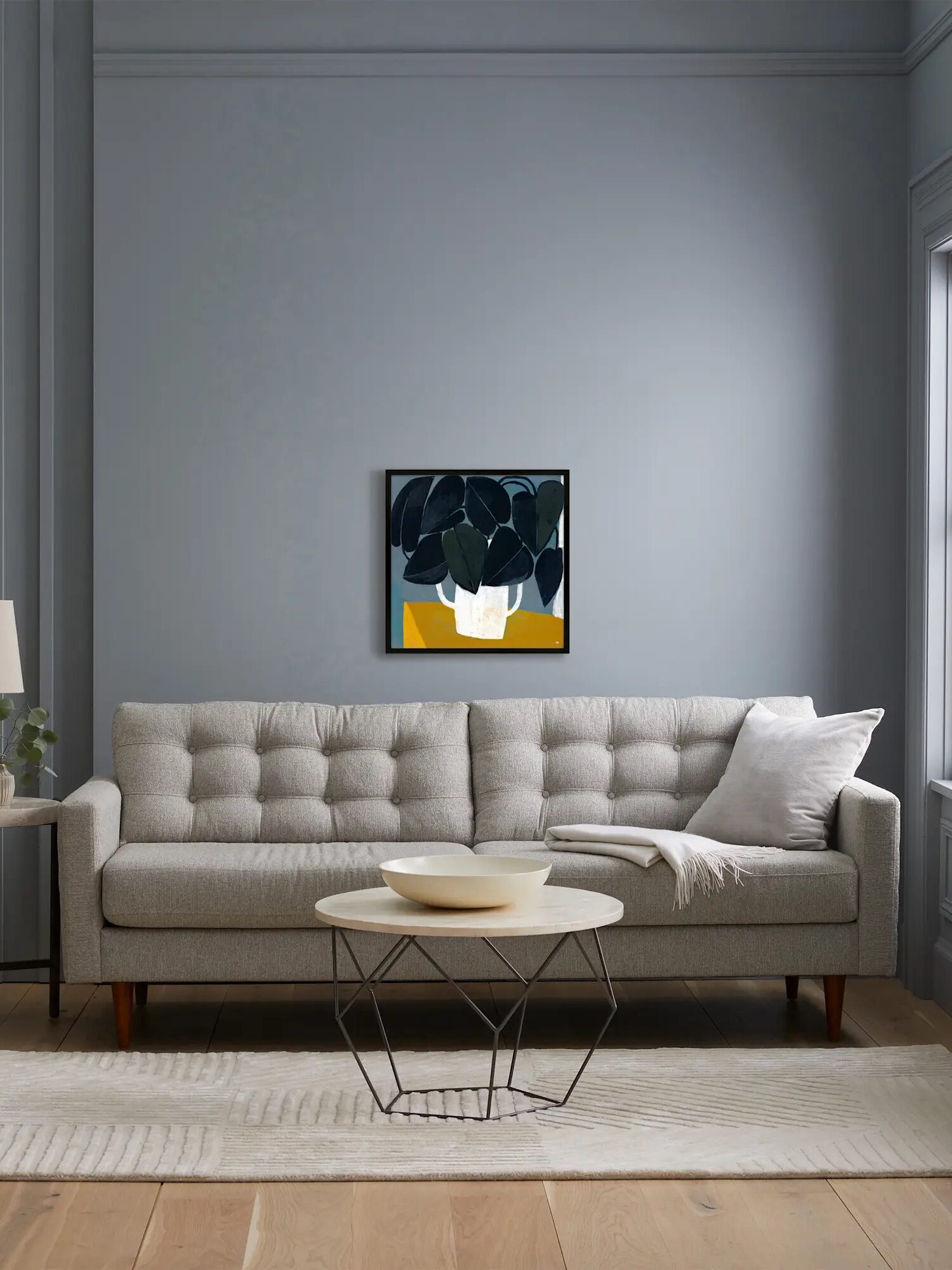 A modern living room with a gray sofa, a geometric coffee table, a rug, and a painting on the wall. A window allows natural light into the space.