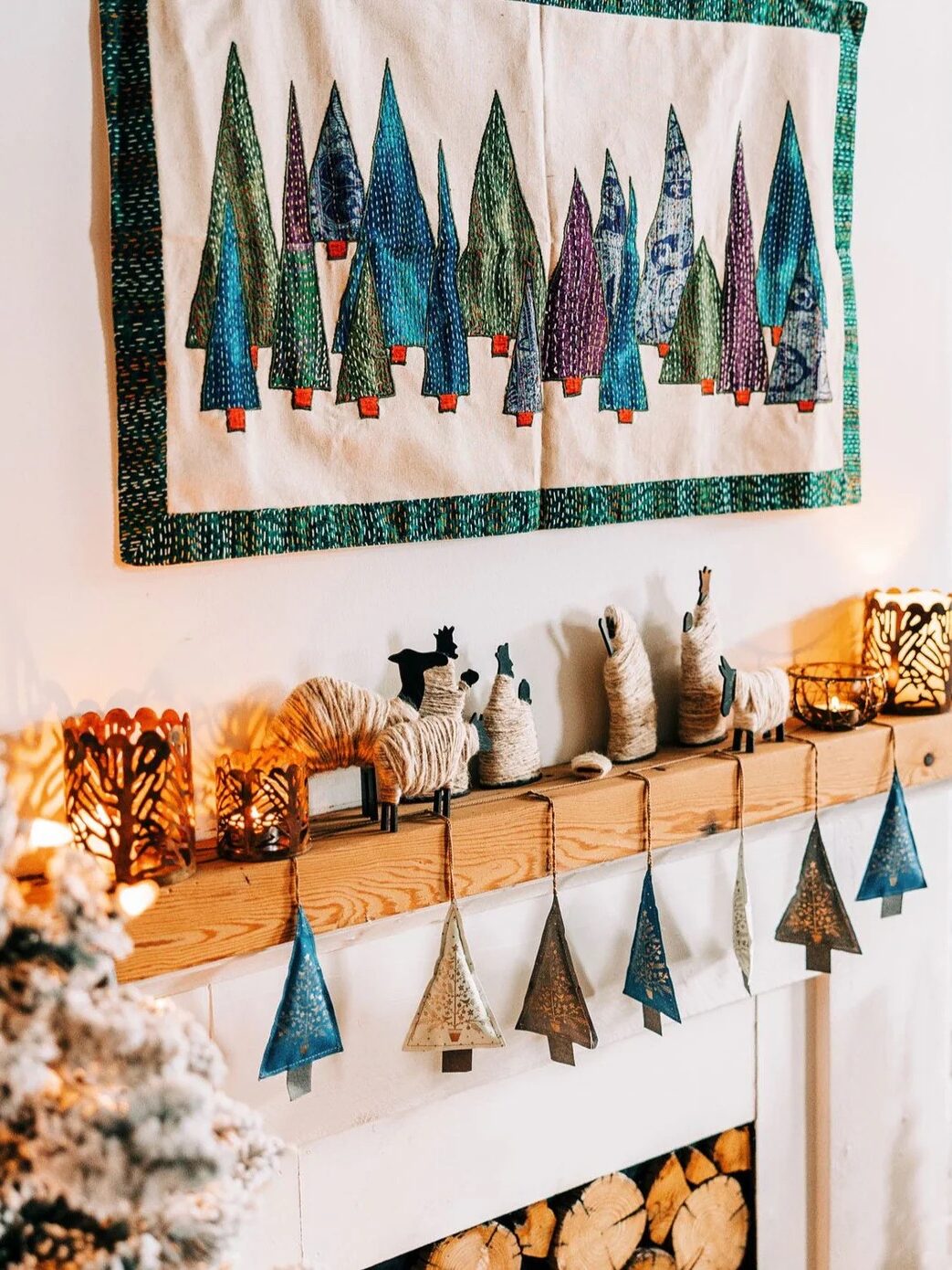 A cozy fireplace mantel with a quilted tree wall hanging above, decorated with candles and ornaments. A small tree and lit candles add warmth to the scene.