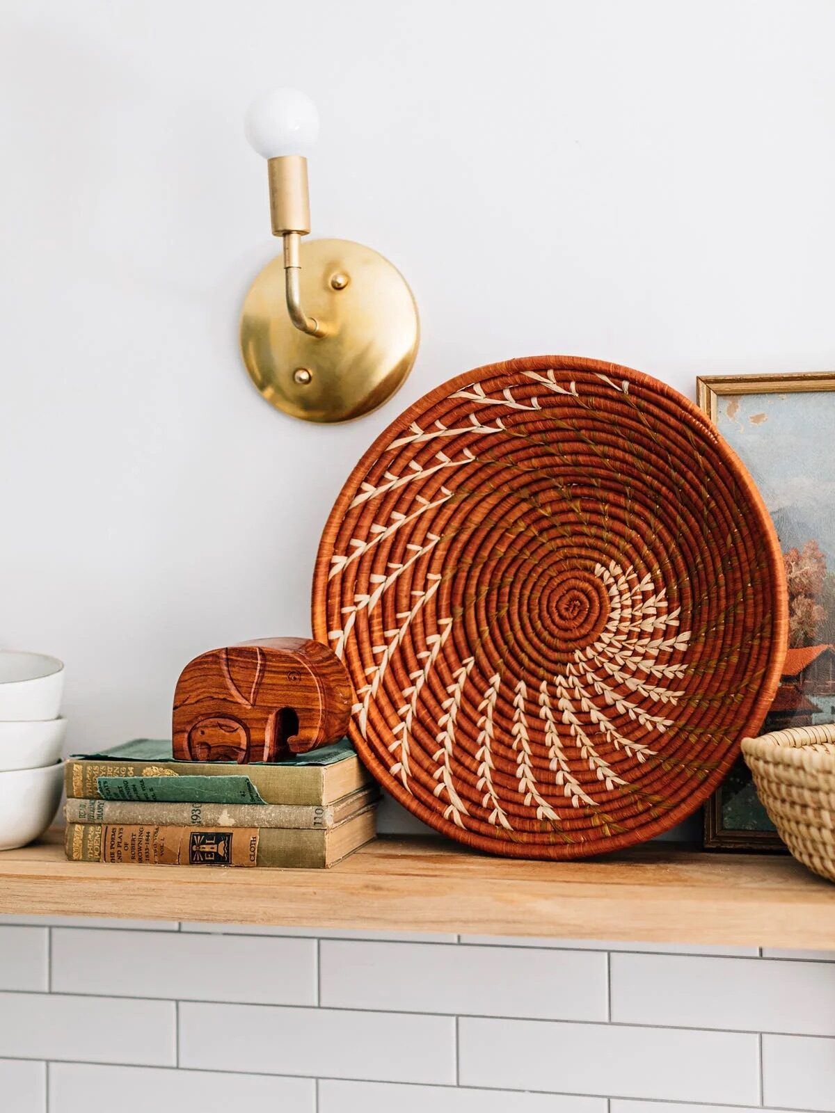 Woven basket, books, wooden elephant, and painting on a shelf under a brass wall lamp with a round bulb.