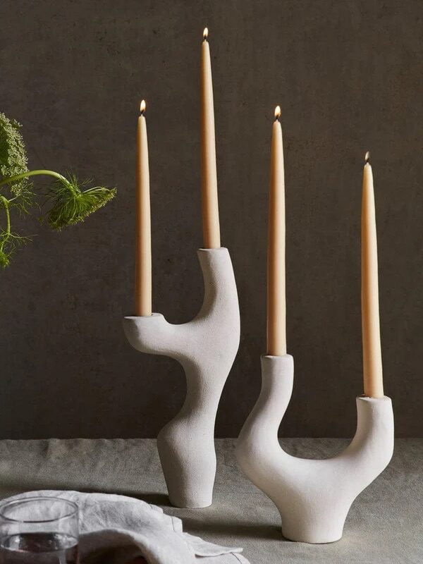 Three lit candles in sculptural ceramic holders are on a table with a vase holding greenery and two glasses nearby.