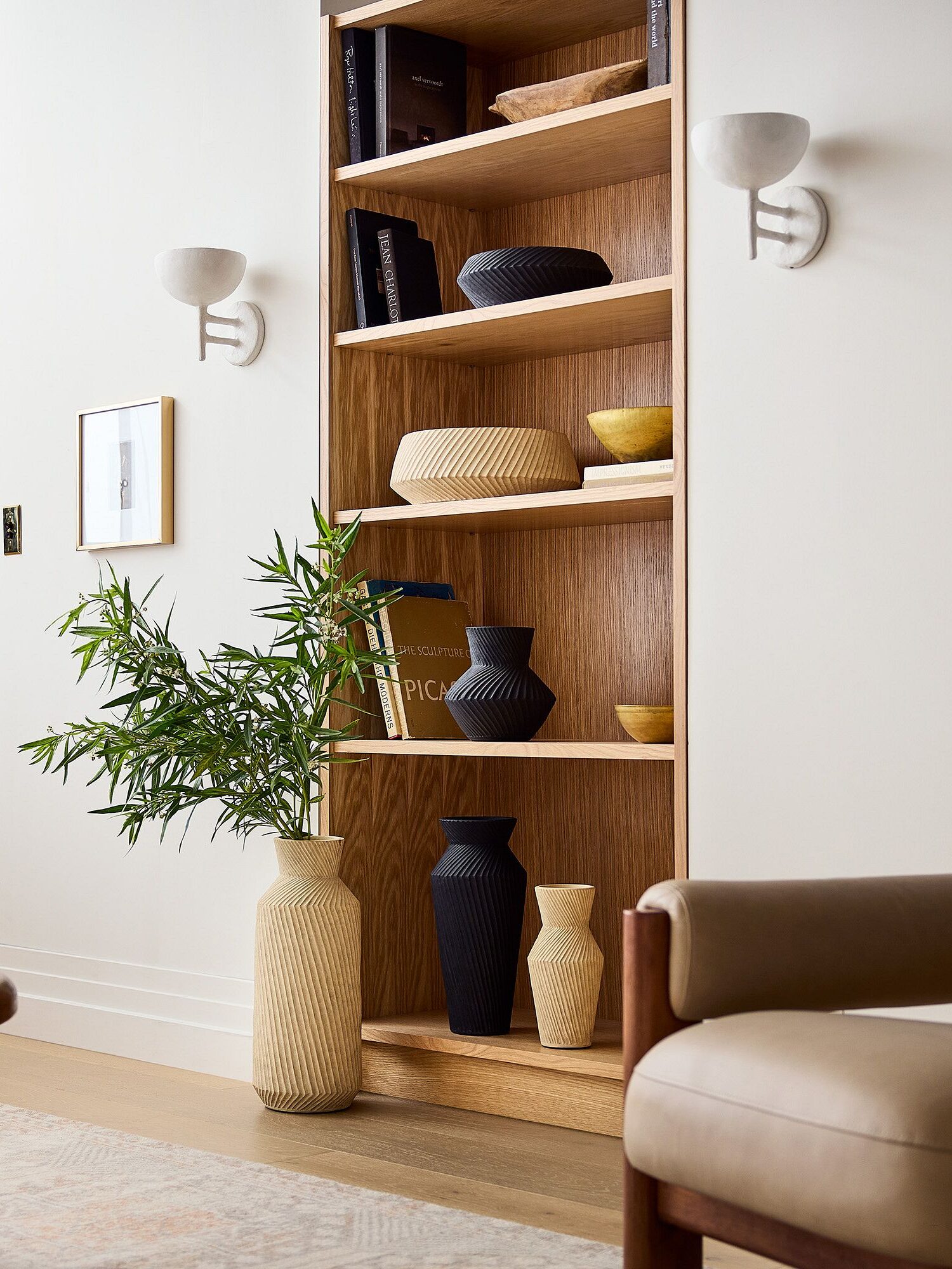 Modern living room with a wooden bookshelf displaying various vases. Artwork and a lamp are on the walls. A potted plant sits on the floor next to a wooden coffee table and comfortable seating.