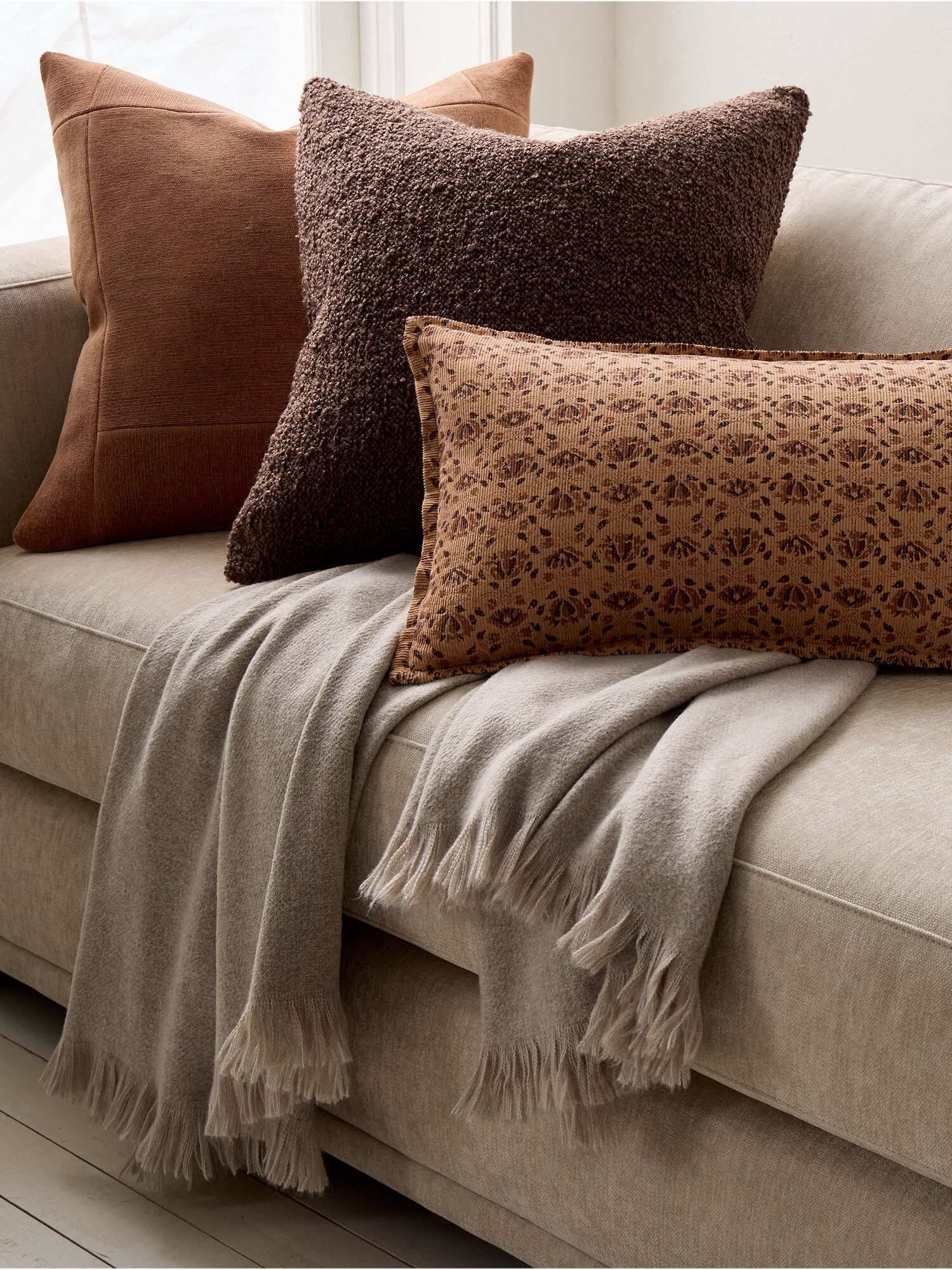 A beige sofa with a gray fringed throw blanket and three decorative pillows in brown tones are arranged on it.