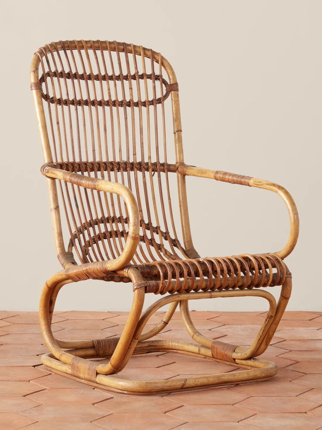 A rattan rocking chair with a curved frame and slatted seat and backrest, placed on a terracotta tile floor against a plain background.