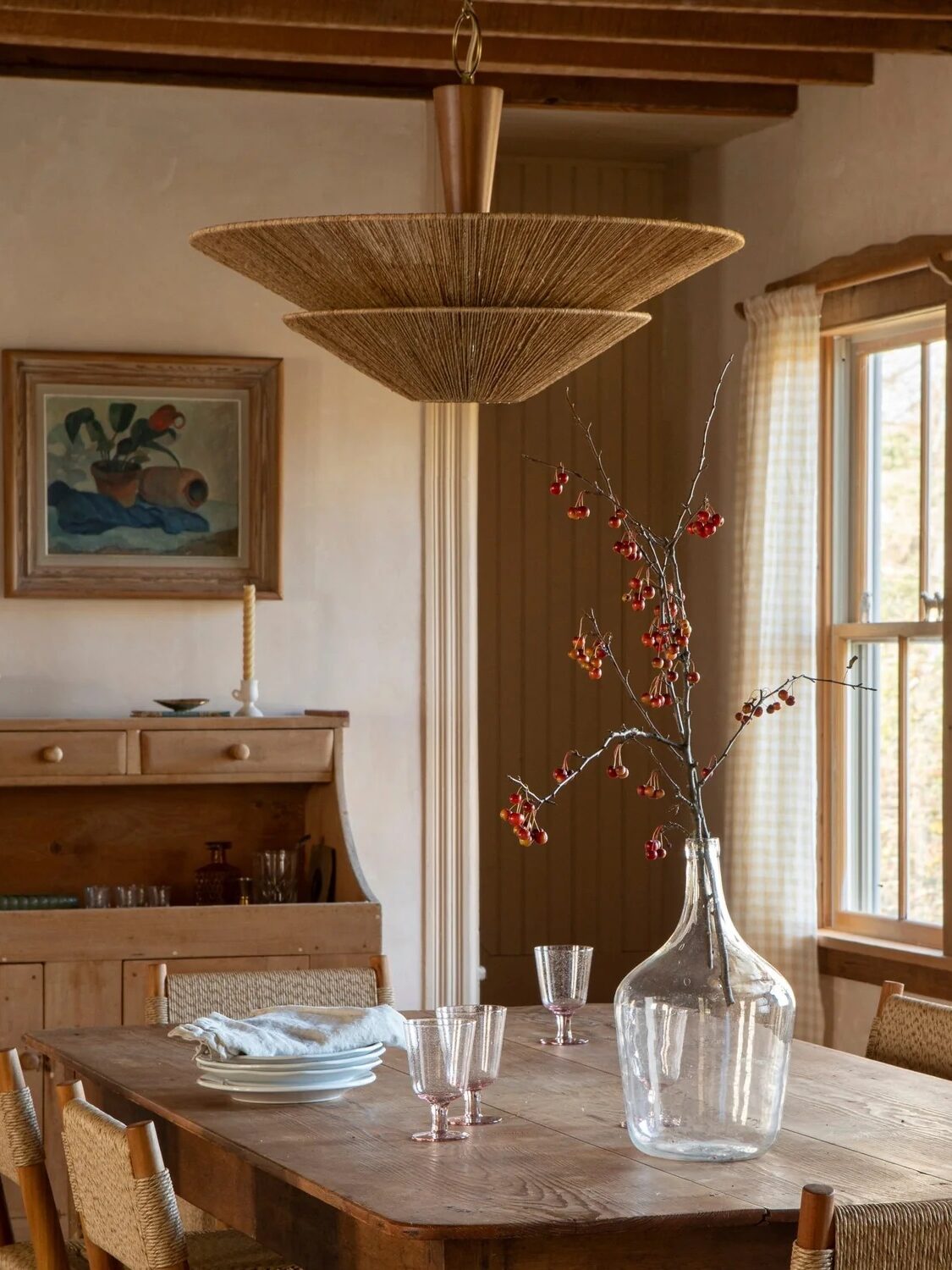 A rustic dining room with a wooden table, chairs, and a glass vase with branches. A large woven light fixture hangs above. Sunlight filters through checked curtains on tall windows.