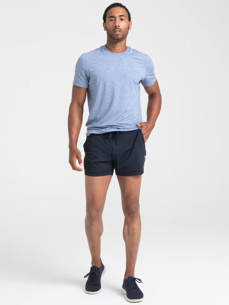 Man wearing a blue t-shirt, black shorts, and black shoes stands against a neutral background.