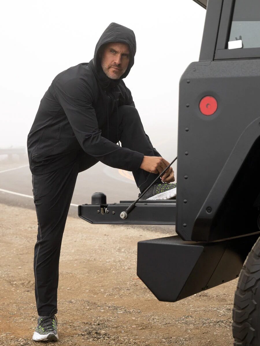 A person wearing a hooded jacket ties their shoe on the back of a black vehicle in a foggy outdoor setting.