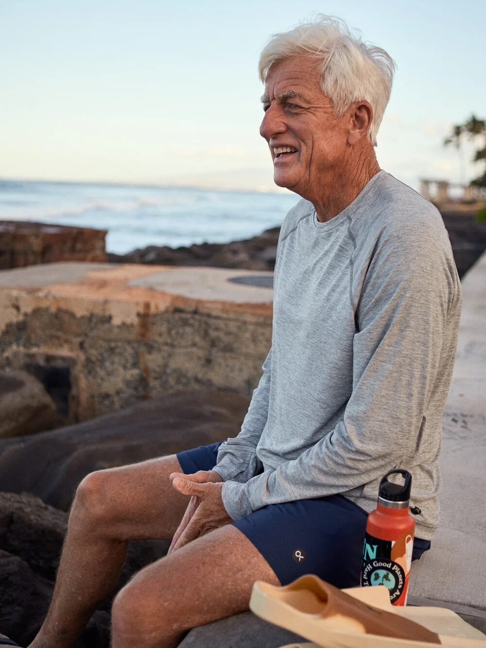 An older man in a gray long-sleeve shirt sits on a rock near the ocean, smiling. A water bottle and a book are beside him.