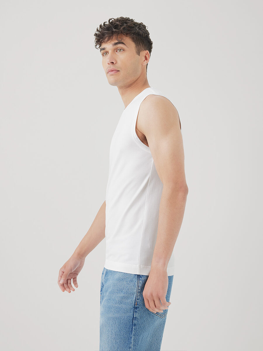 A person wearing a white sleeveless shirt and blue jeans is standing against a plain background, facing slightly to the side.