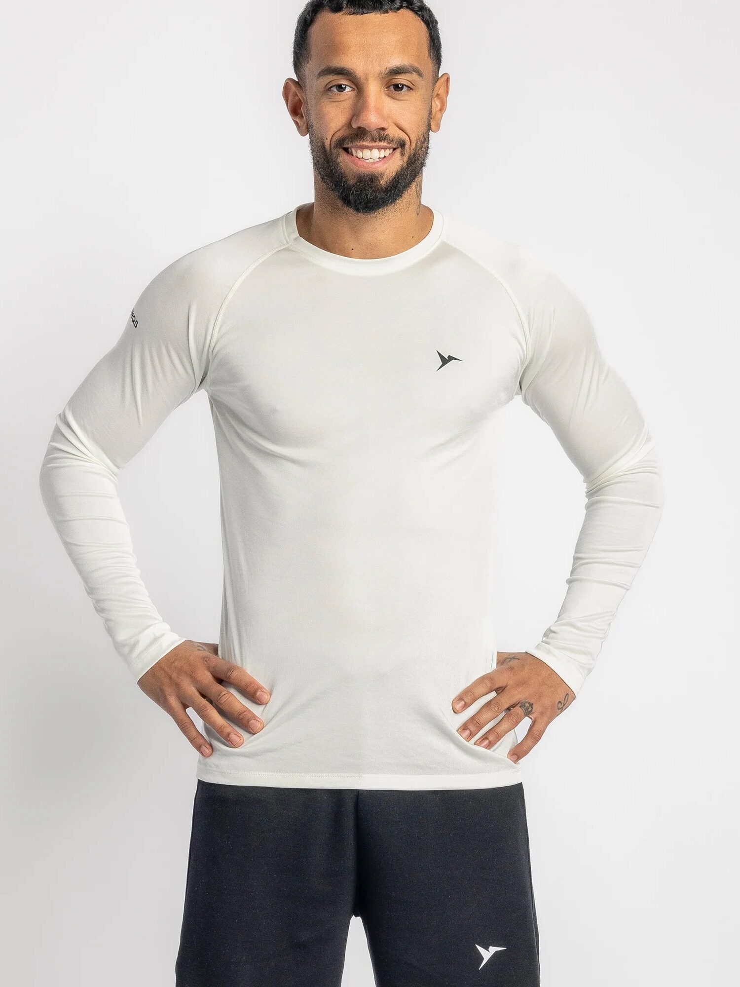 Person wearing a white long-sleeve athletic shirt and black shorts stands against a plain background, smiling with hands on hips.