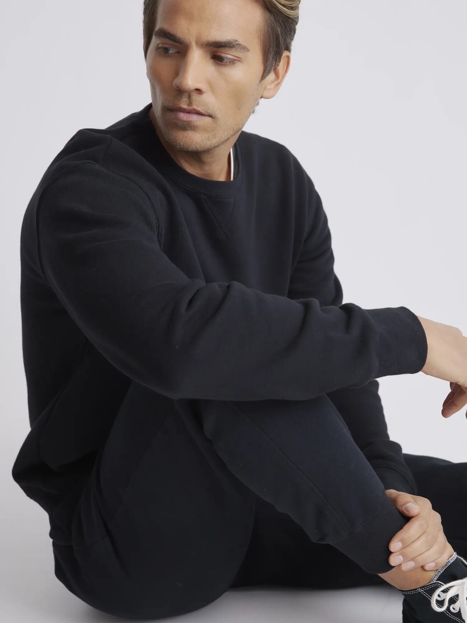 A person sits on the floor wearing a black sweatshirt and pants, looking to the side against a plain white background.