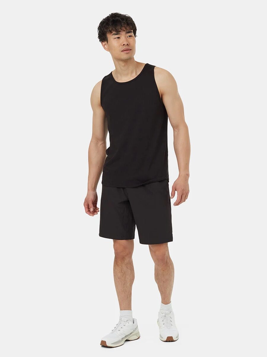 Man wearing a black tank top, black shorts, and white sneakers stands against a plain white background.