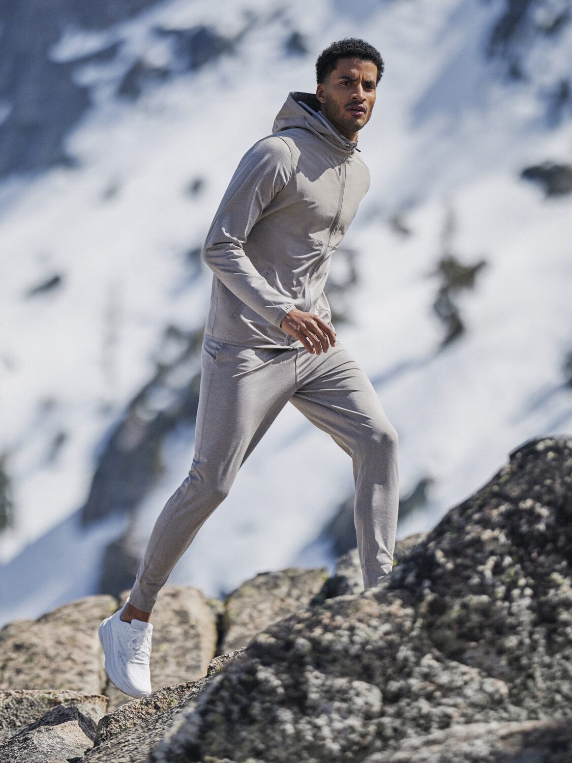 Man in a light gray tracksuit hikes on rocky terrain with snow-covered mountains in the background.