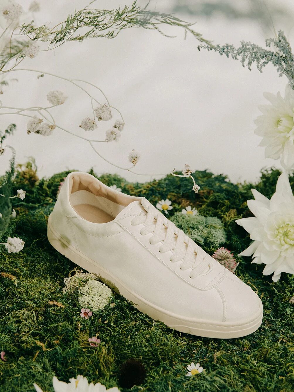 A single white sneaker on green moss surrounded by white flowers and greenery.