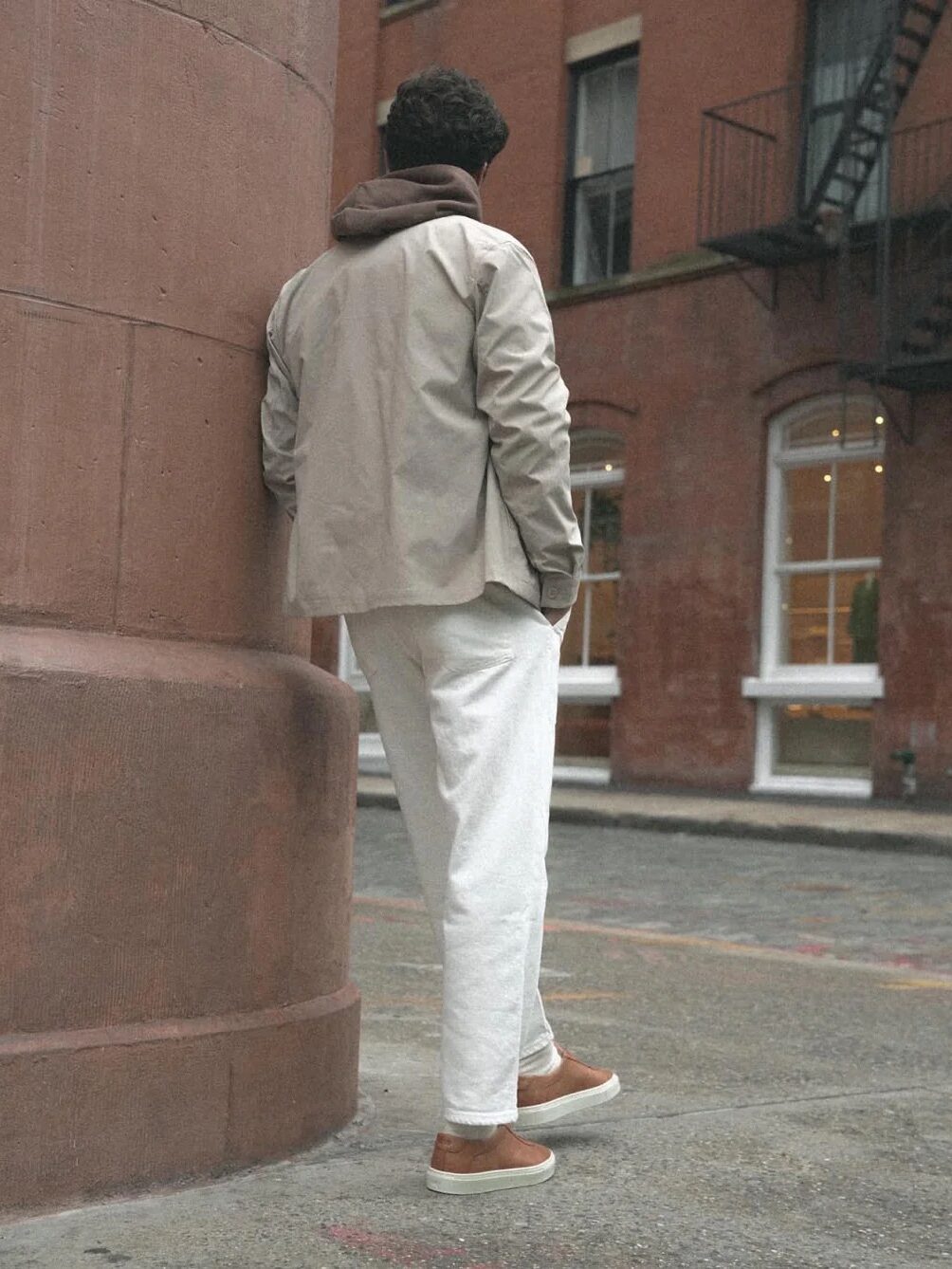 A person in a beige jacket and white pants leans against a brick building on a city street, facing away.