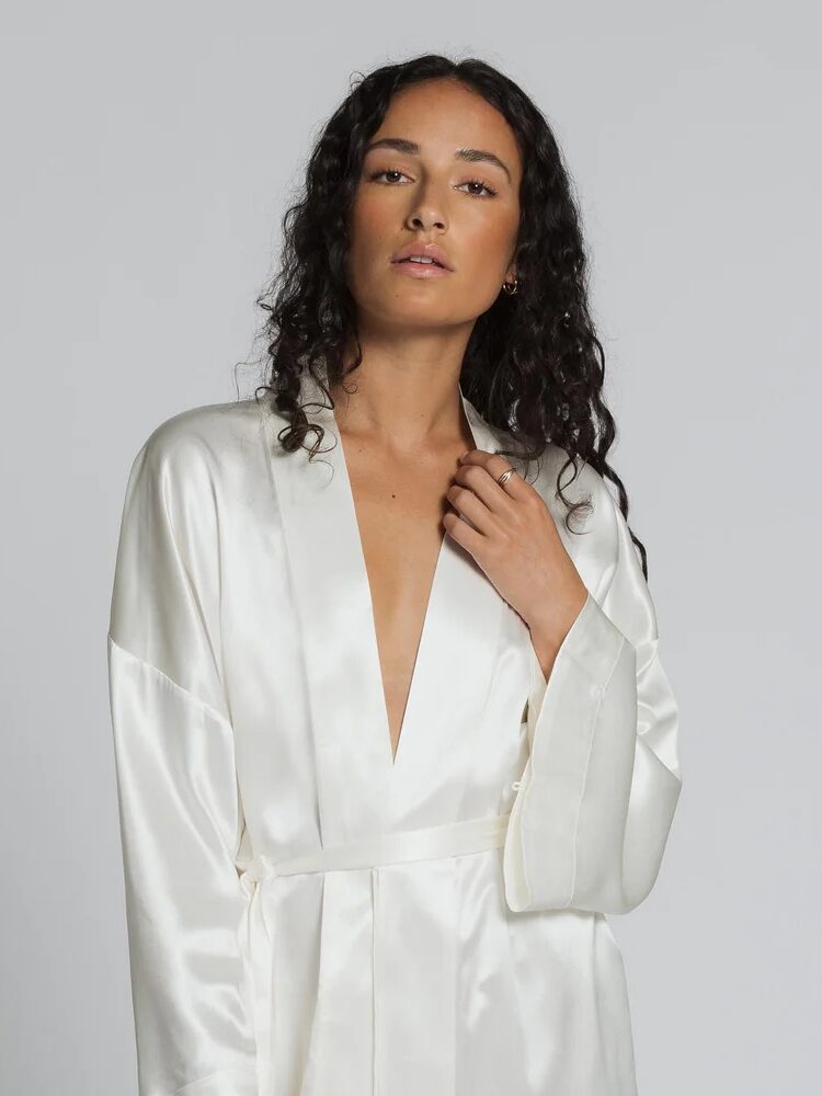 A person with long curly hair wears a silky white robe against a plain background.