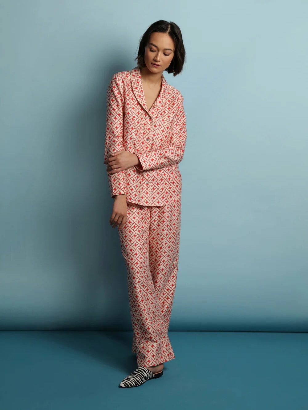 A person wearing a patterned pajama set and striped shoes stands against a light blue background. They have dark hair and are resting one arm across their body.