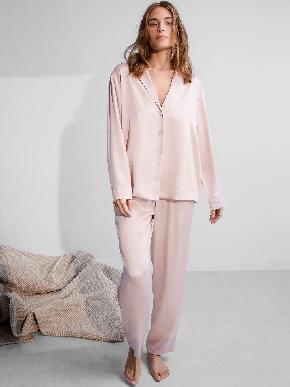 Woman wearing a light pink pajama set stands barefoot in a minimally decorated room with a textured blanket on the floor nearby.