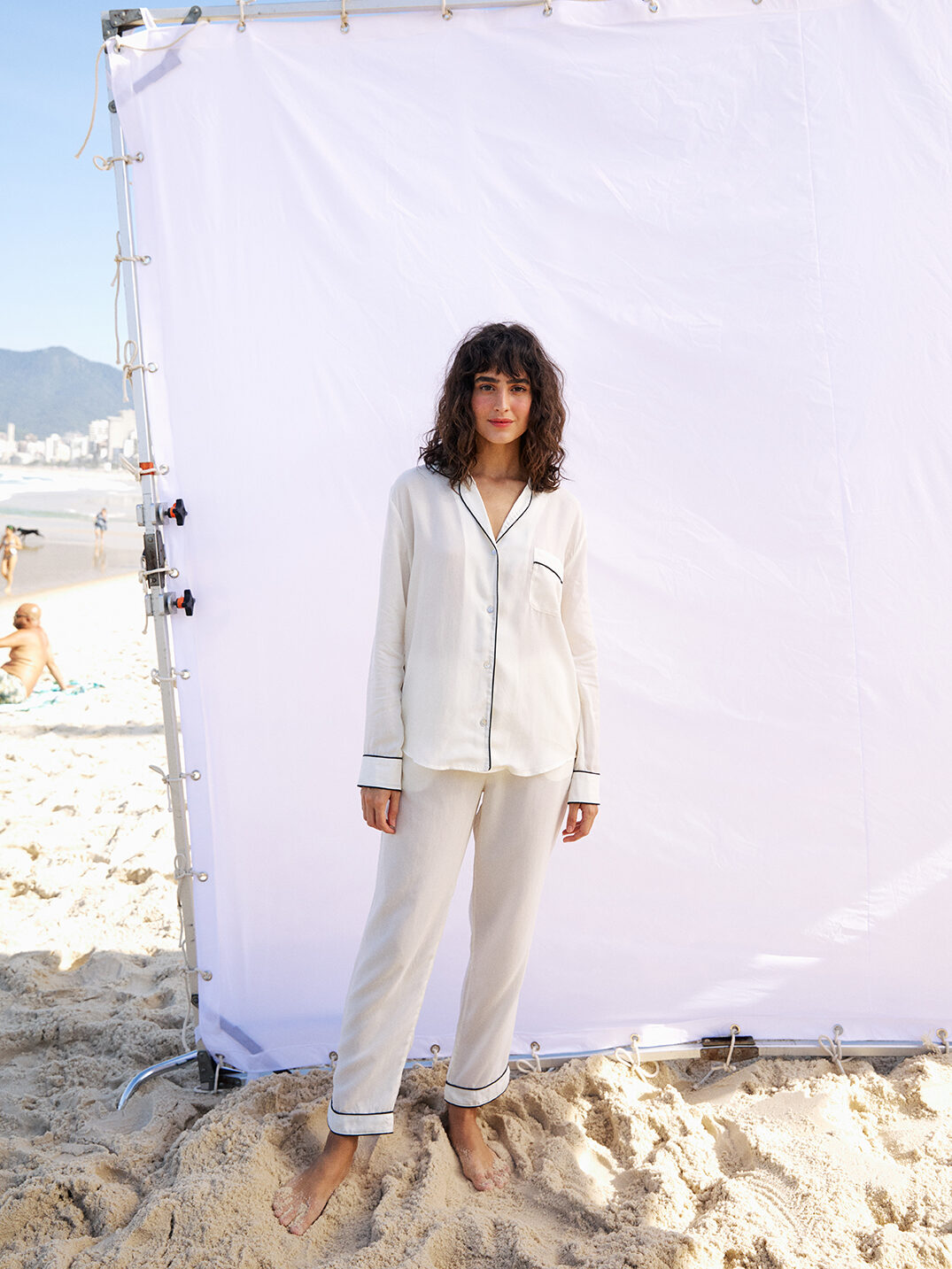 A model in front of a white photo backdrop on a beach wears  white silk pajamas with black piping