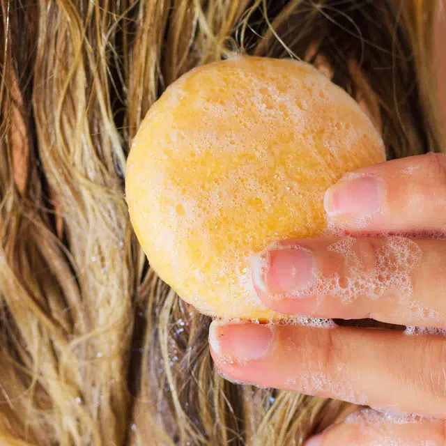 Close-up of a person washing their hair with a foamy, round yellow soap. Fingers are visible holding the soap against damp hair.