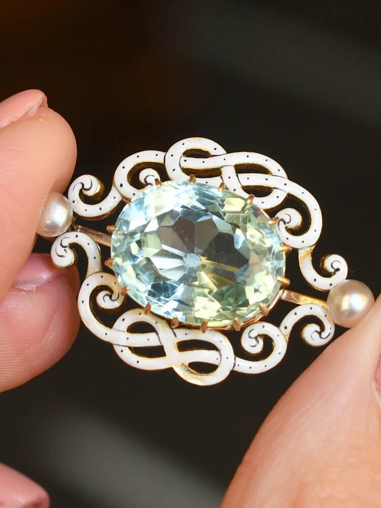 Close-up of hands holding an ornate brooch featuring an oval blue gemstone, intricate white enamel design, and two small pearls on either side.