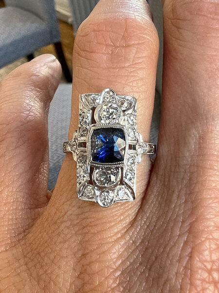 A hand displays an ornate silver ring featuring a large blue gemstone surrounded by intricate detailing and smaller clear stones.