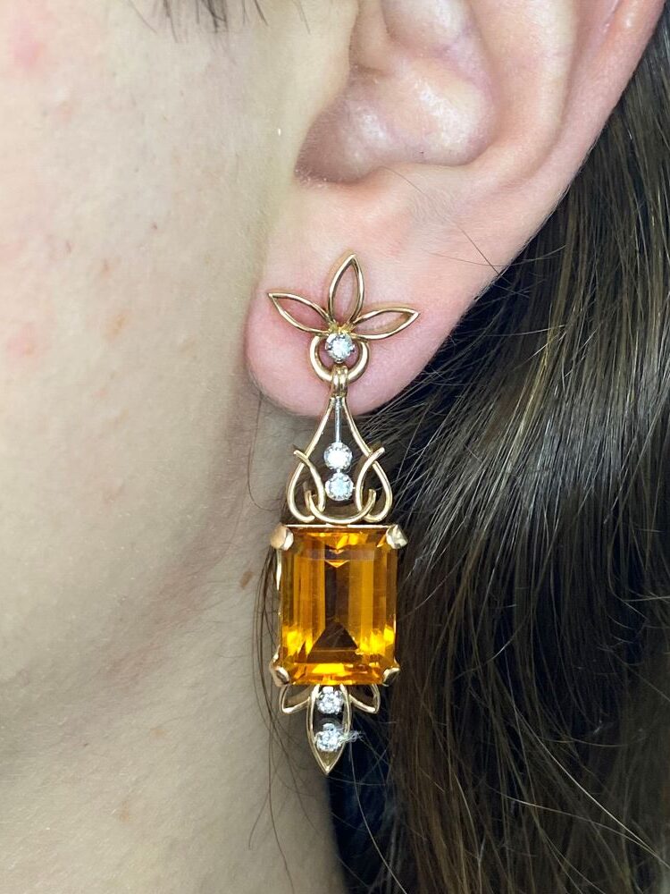 A close-up of a person's ear wearing a gold earring with a large rectangular amber gem, small diamonds, and floral design elements.