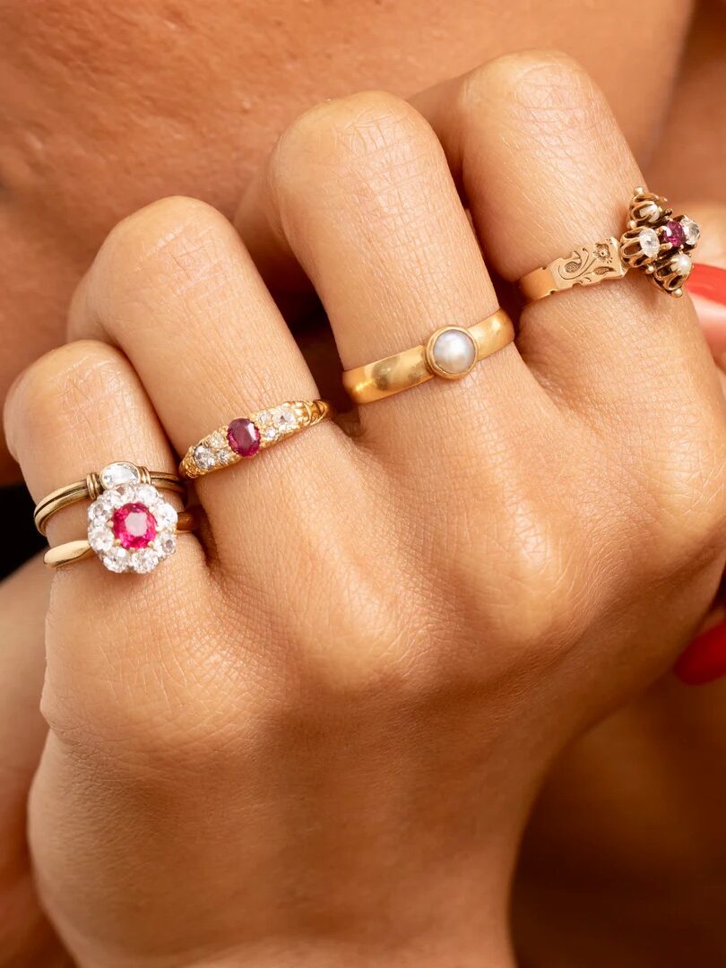 Close-up of a hand displaying four gold rings with various gemstones on the fingers, and nails painted in a bright orange-red color.