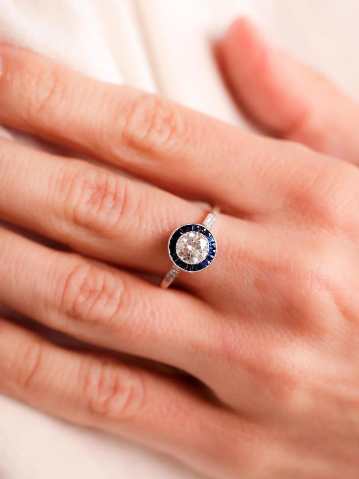 A hand wearing a ring with a large central jewel surrounded by smaller blue stones on a silver band.