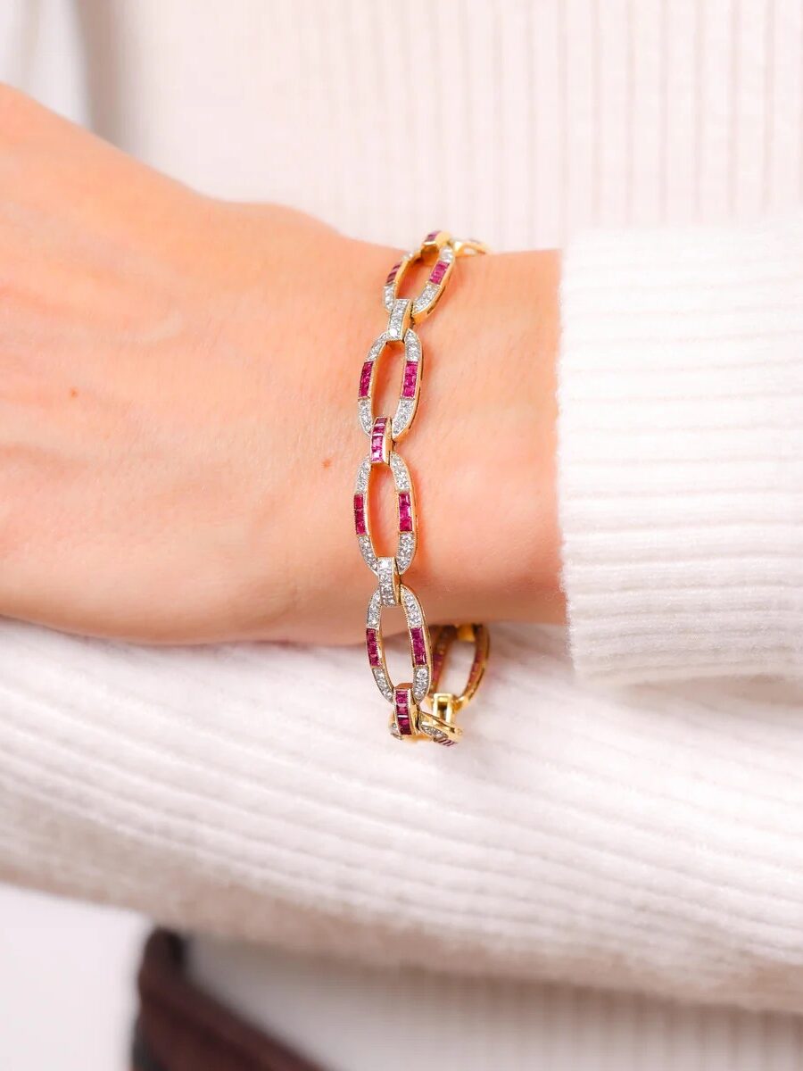 A woman's arm wearing a gold chain bracelet featuring oval red gemstones and small clear stones. She is dressed in a white ribbed sweater.