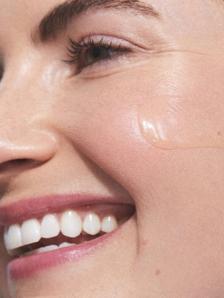 Close-up of a person smiling with a visible glossy skincare product applied on their cheek.