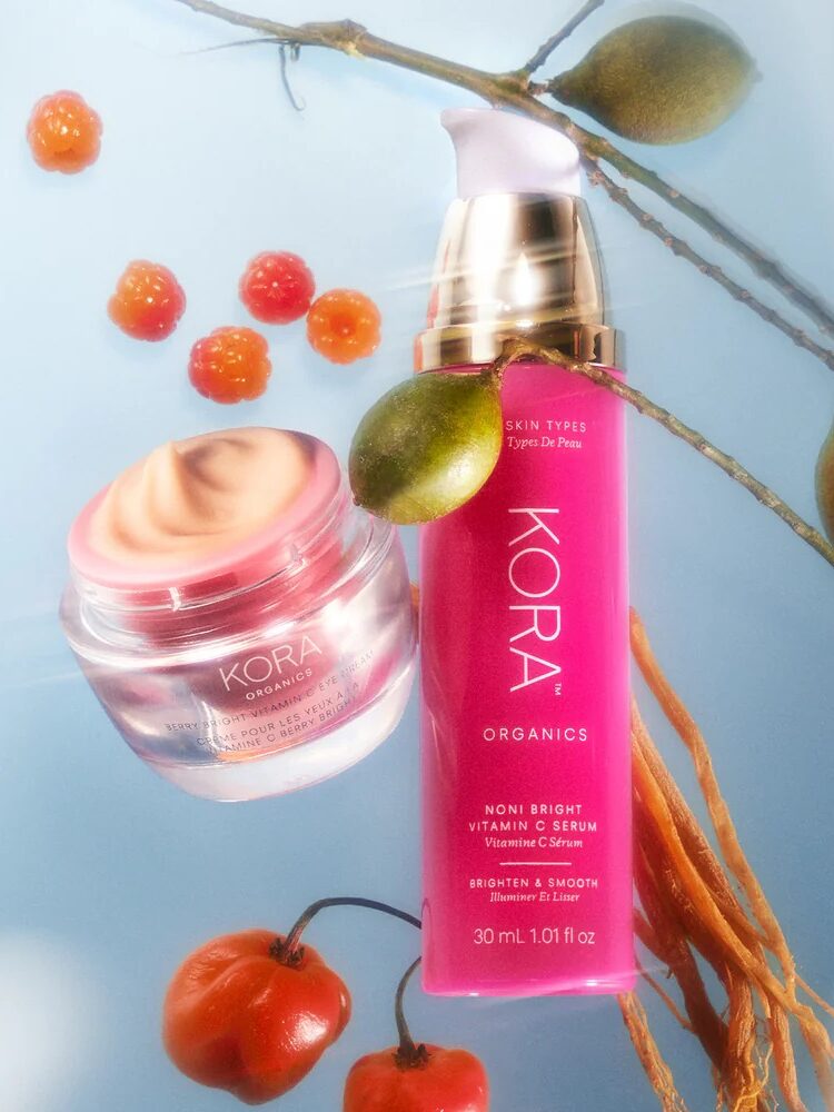 A pink bottle and jar of Kora Organics skincare products are placed among fruits and branches on a light background.