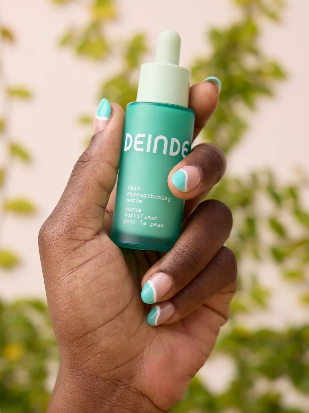 Hand holding a green bottle labeled "DEINDE skin-strengthening serum," with green and white manicured nails. A blurred leafy background is visible.