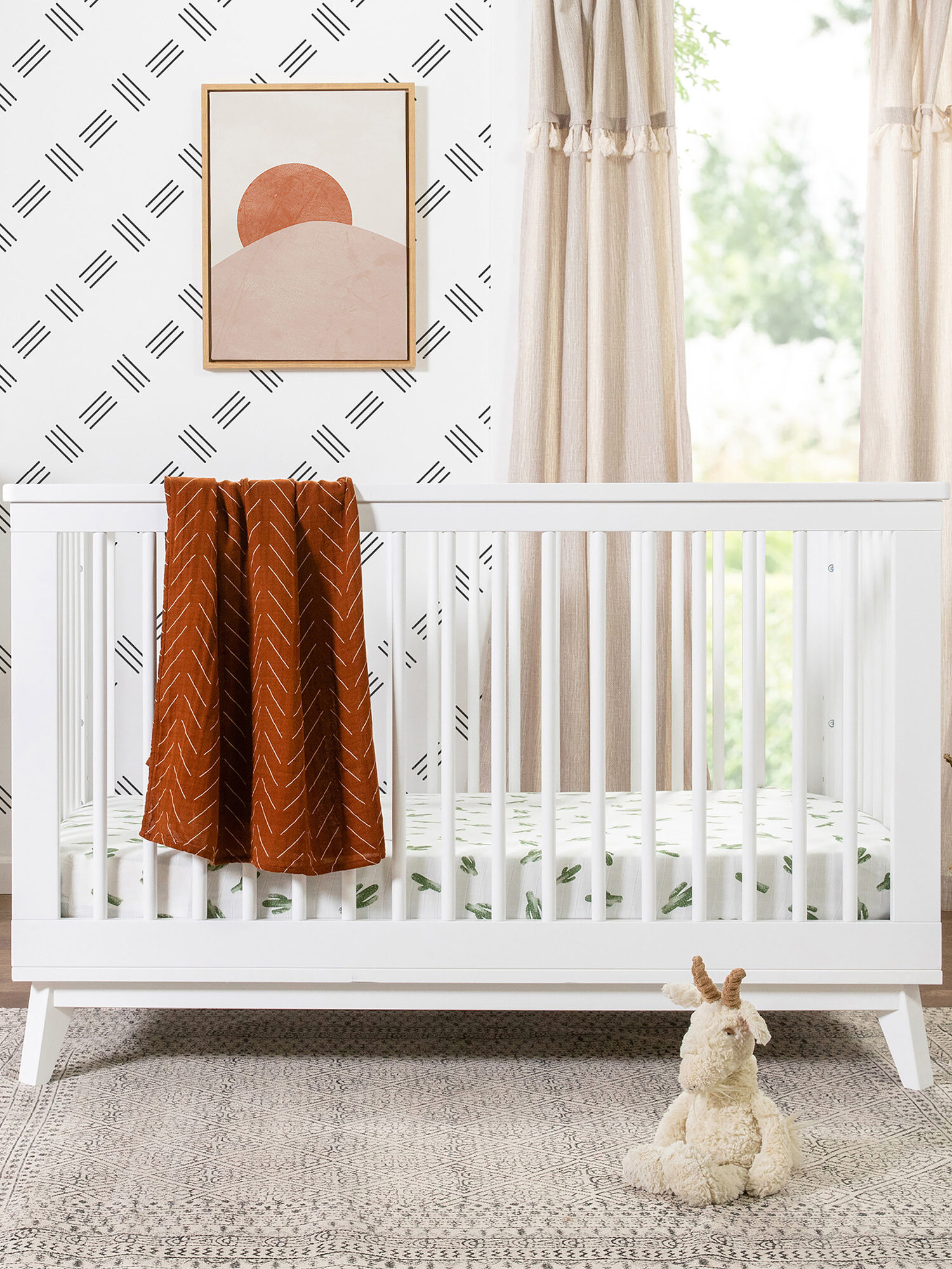 A white baby crib with a rust-colored blanket draped over the side is set against a patterned wall. There is a small stuffed animal on the floor and a plant nearby.