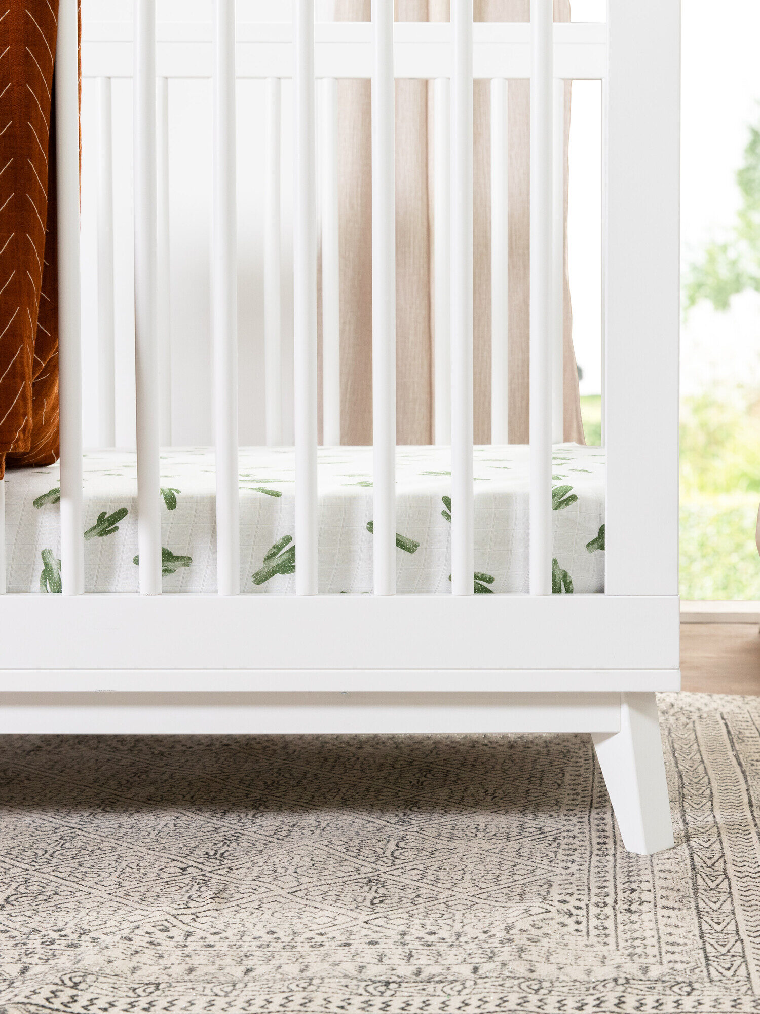 A white crib with a cactus-patterned sheet is next to a tall potted plant on a woven rug.