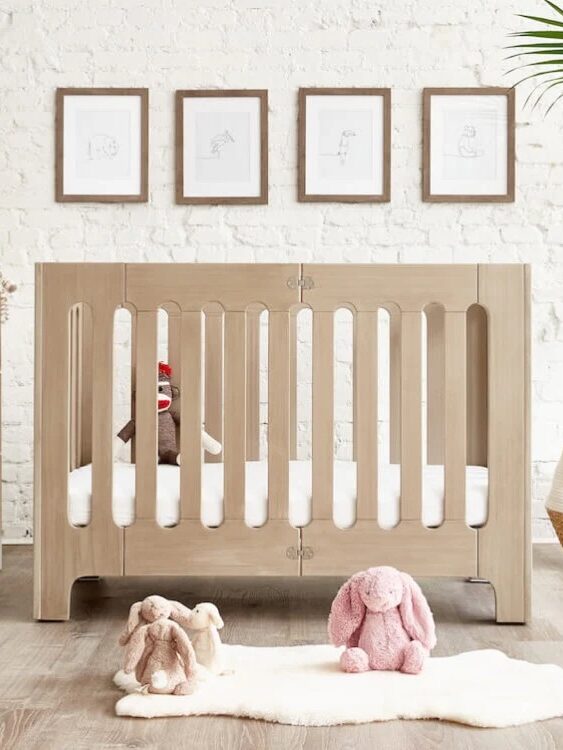 A wooden crib with stuffed animals inside and outside sits in a nursery. White brick wall with framed pictures in the background, and a large plant on the right.