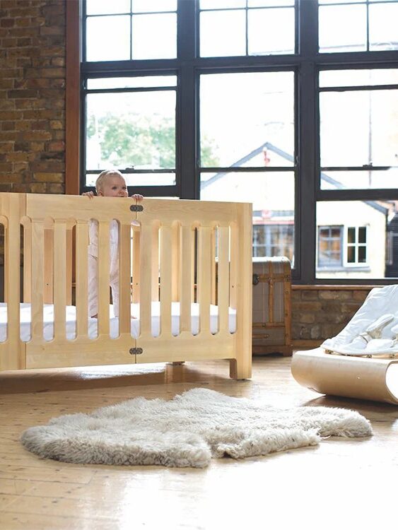 A baby stands in a wooden crib beside a soft rug and a chair in a room with large windows and exposed brick walls.