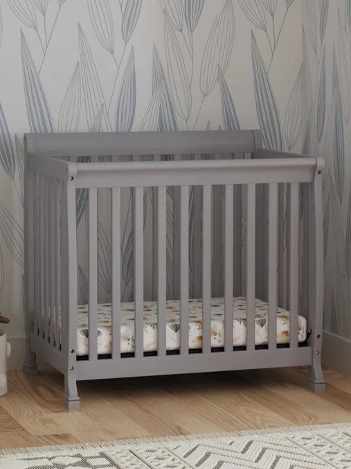 Gray crib with slatted sides and a patterned mattress inside, set against a patterned wallpaper background with a rug on the wooden floor.